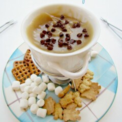 Cup full of cookie dough fondue on a plaid plate with marshmallows, pretzels and teddy grahams on plate
