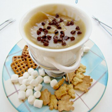 Cup full of cookie dough fondue on a plaid plate with marshmallows, pretzels and teddy grahams on plate