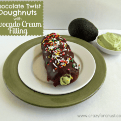 Chocolate twist doughnuts with avocado cream filling on a white plate, on top of a green charger, with a bowl of avocado cream, plus an avocado in the background.