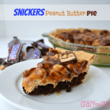 A whole snickers pie with one slice on a white plate and opened snickers candy bar wrappers on the side.