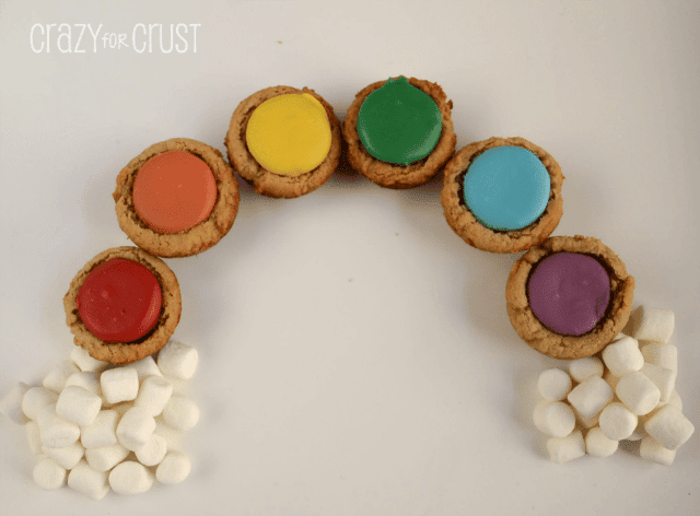 peanut butter cookies cups with rainbow colored candies in the center in the shape of a rainbow with marshmallow clouds