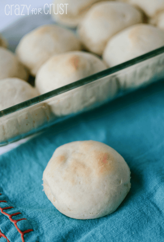 One dinner roll on turquoise cloth napkin with full glass dish and more rolls in the background