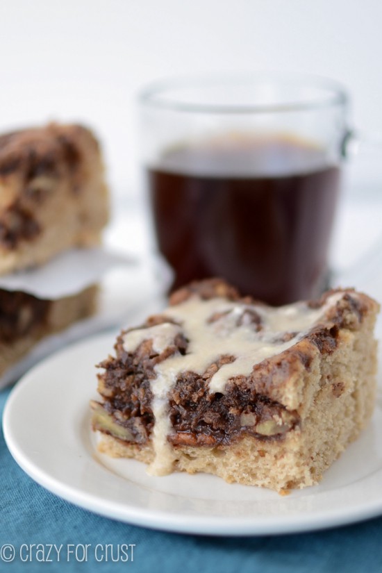 Close up photo of Mocha Coffee Cake piece with icing dripping off on a white plate with a cup of coffee in the background