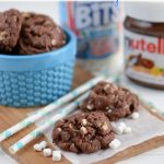 Rocky road cookies on a wood cutting board with straws and ingredients in the background