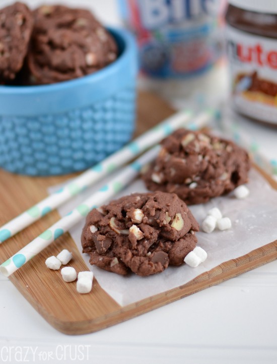 Rocky Road Nutella Pudding Cookies on wood cutting board with ingredients behind and marshmallows and straws next to the cookies