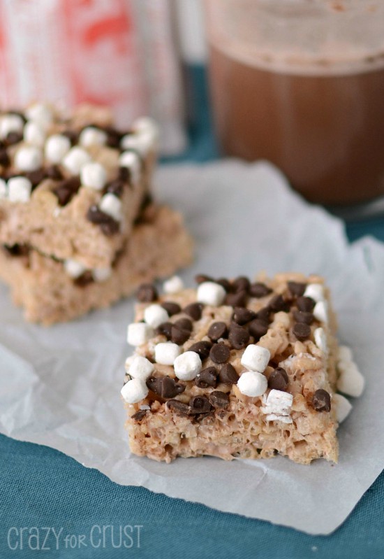 Hot Chocolate Krispie Treats with marshmallows and chocolate chips on parchment paper with hot cocoa in back