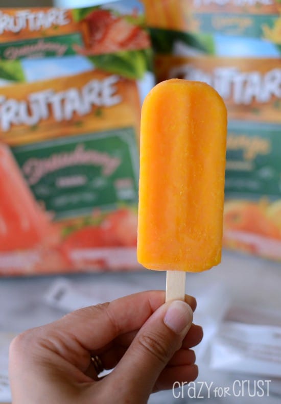 Hand holding a orange popsicle with popsicle boxes out of focus in the background