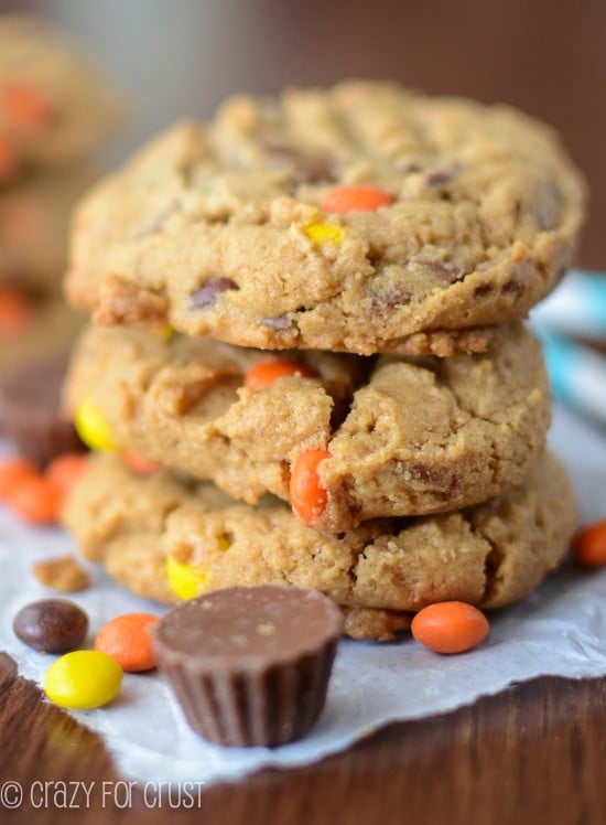 Stack of three Flourless Reese's Explosion Peanut Butter Cookies on a wooden cutting board with peanut butter candies around them