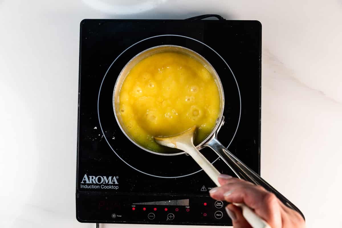 La main remue un bol de liquide jaune, probablement des œufs, avec une louche sur une cuisinière à induction aromatique. Le pot à bulles est posé sur une surface blanche et les ingrédients d'un délicieux gâteau au chocolat allemand sont probablement en cours de préparation.