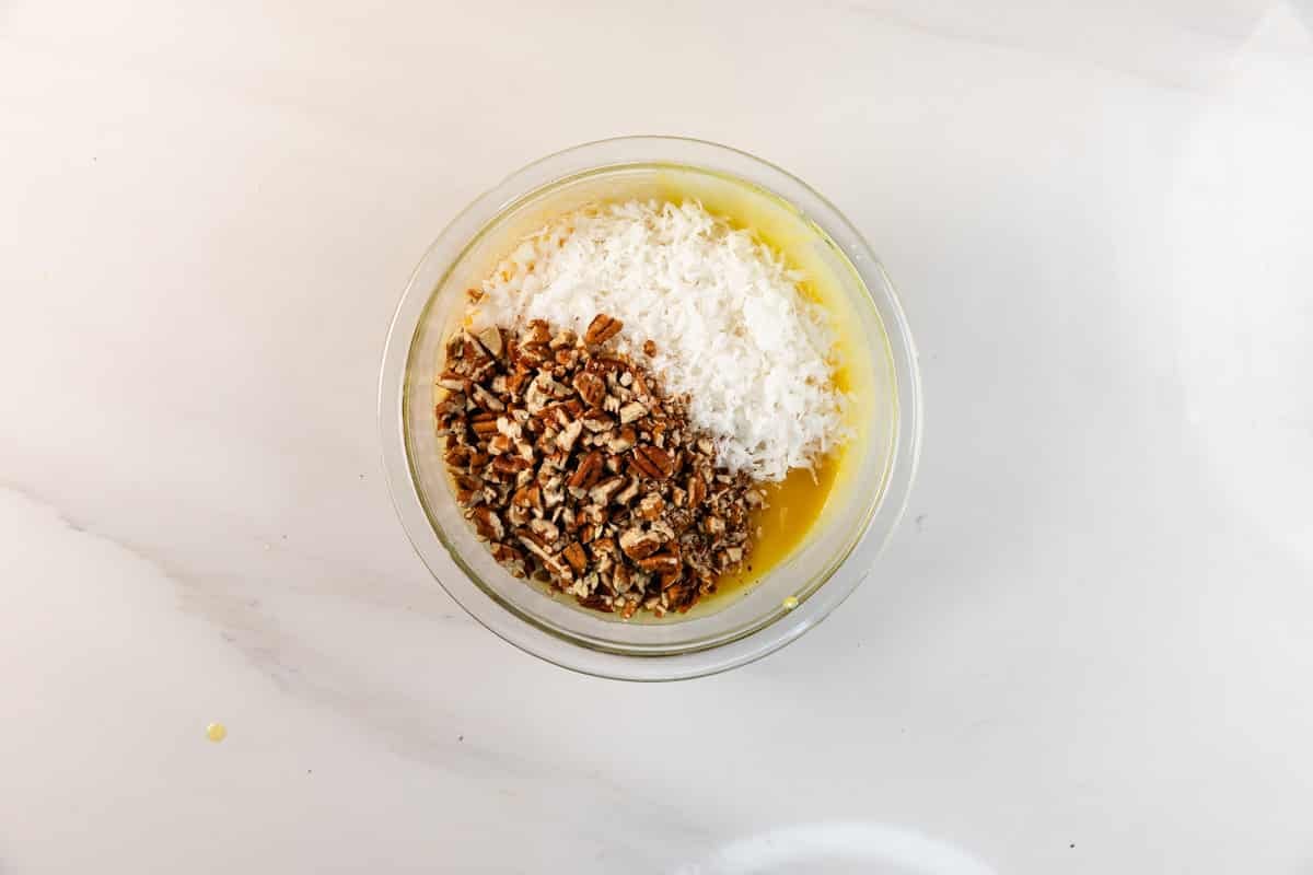Un bol en verre rempli de beurre fondu, de noix de coco râpée et de pacanes hachées – les ingrédients classiques du gâteau au chocolat allemand – repose sur un dessus de marbre blanc.