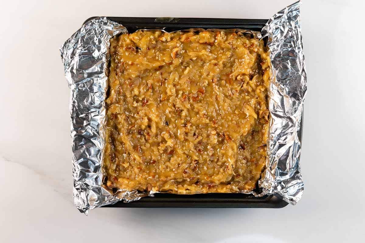 Un plat de cuisson carré recouvert de papier d'aluminium froissé contient une couche de pacanes et de noix de coco, uniformément répartie sur la surface de ces brownies au chocolat allemands décadents.