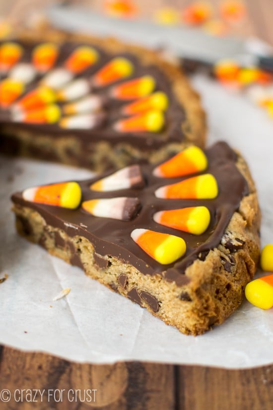 Closeup shot of a slice of Harvest Chocolate Chip Cookie Cake in front of whole cookie cake on white parchment paper