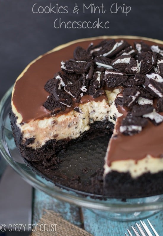 Close up shot of Cookies and Mint Chip Cheesecake with one slice missing on clear cake stand with recipe title in text on top of photo