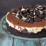 cheesecake on clear cake stand with oreo crust and chocolate topping and chopped oreos on top
