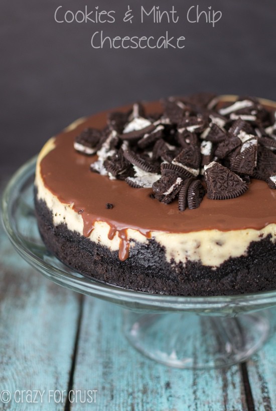 cheesecake on clear cake stand with oreo crust and chocolate topping and chopped oreos on top