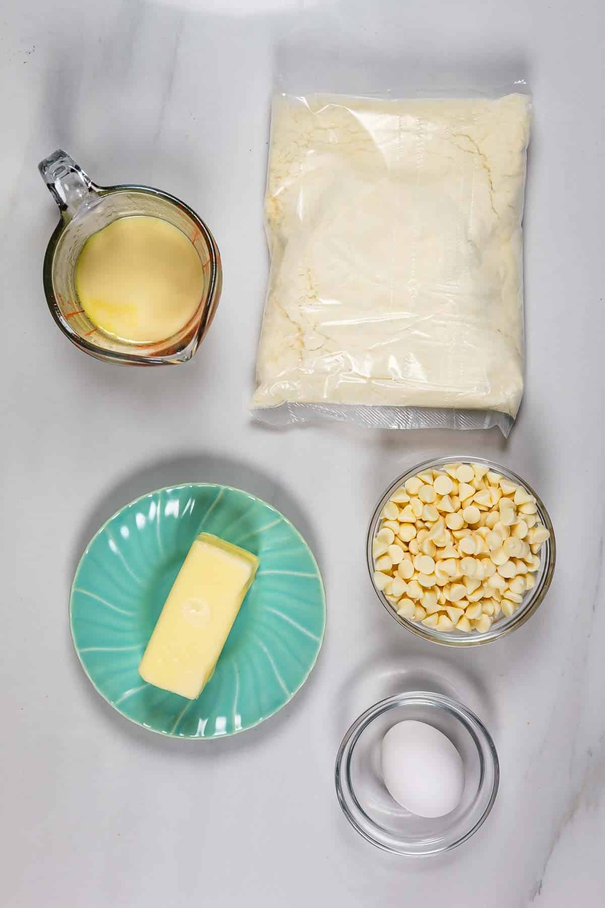 Top-down view of five baking ingredients for Lemon Gooey Bars on a white surface: a measuring cup of liquid, a bag of flour, white chocolate chips in a bowl, a stick of butter on a green plate, and an egg in a small bowl.