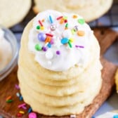 A stack of Perfect Sugar Cookies topped with colorful sprinkles sits on a wooden board, surrounded by more cookies and scattered sprinkles.
