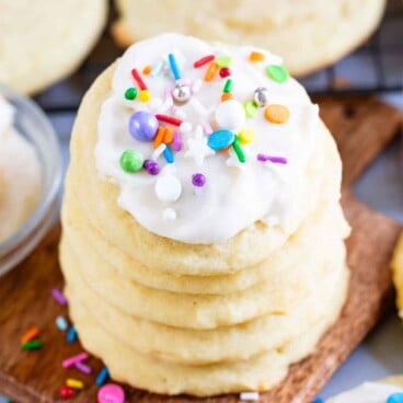 A stack of Perfect Sugar Cookies topped with colorful sprinkles sits on a wooden board, surrounded by more cookies and scattered sprinkles.