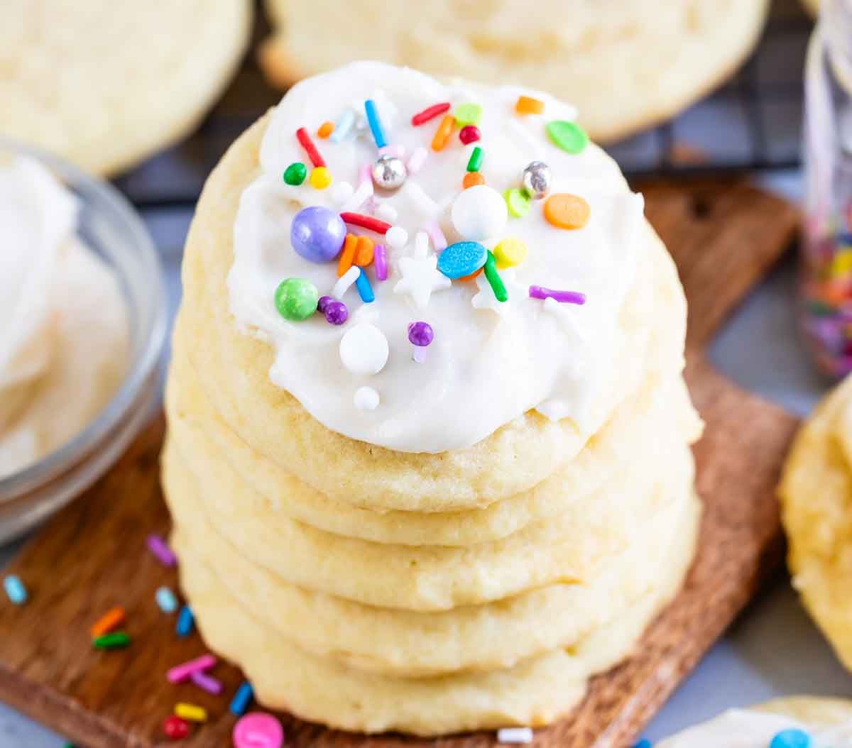 A stack of Perfect Sugar Cookies topped with colorful sprinkles sits on a wooden board, surrounded by more cookies and scattered sprinkles.