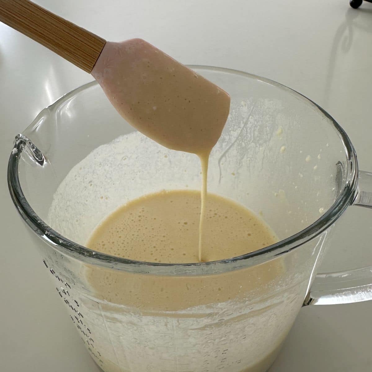Rouleaux de tarte au sucre et à la cannelle - Fous de croûte 12 Une spatule en silicone rose clair prélève une cuillerée de pâte à crêpes brun clair dans une tasse à mesurer en verre transparent remplie de pâte.