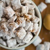 Peanut Butter Snickerdoodle Muddy Buddies sitting in a bowl and all over the table with spoonful of peanut butter laying there