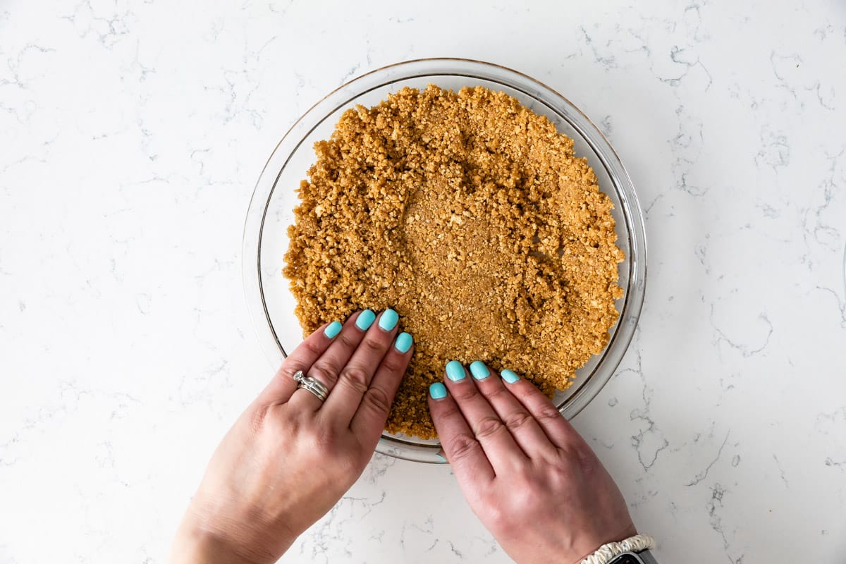 glass pie plate with graham cracker crust crumbs being pressed by hands.