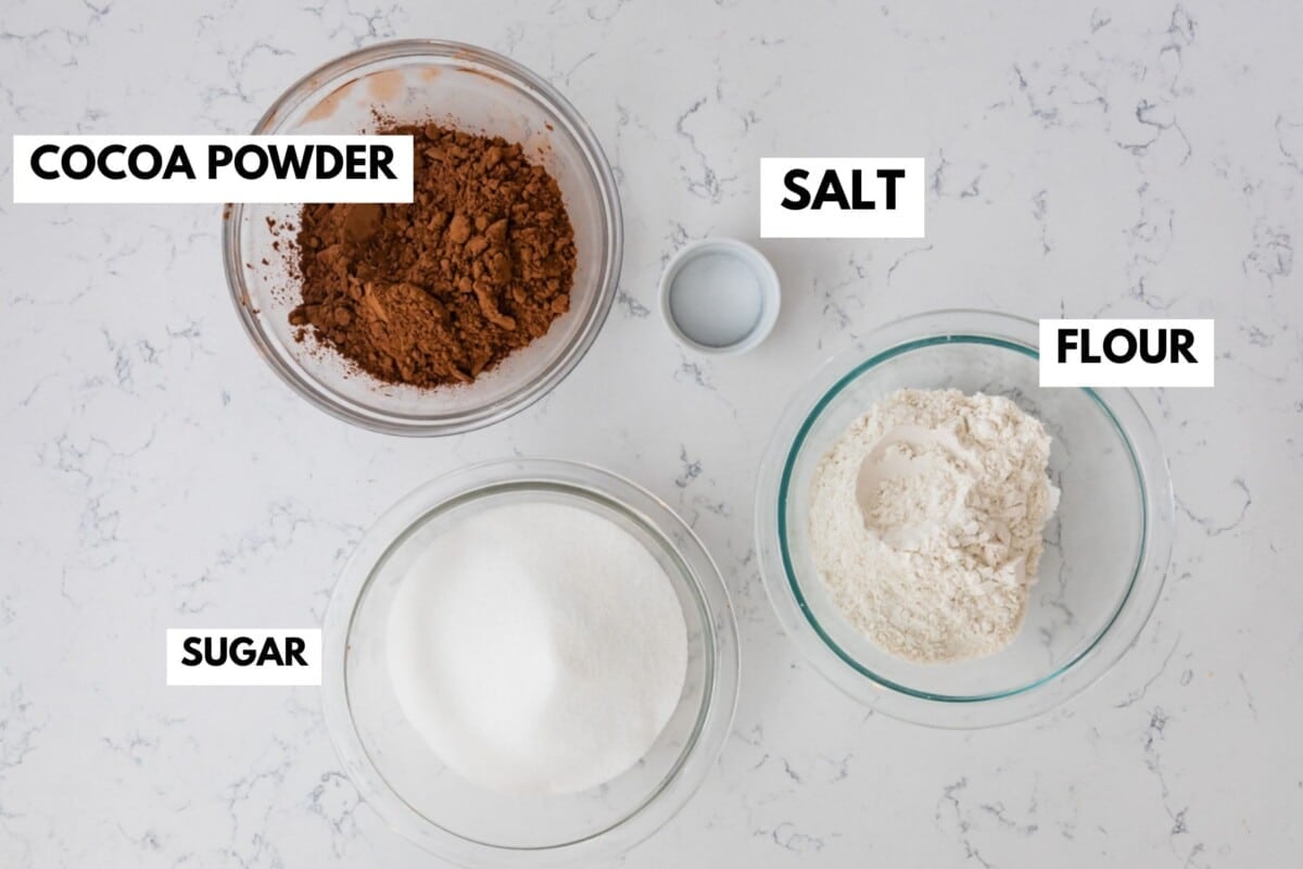 Four labeled bowls on a white surface display the essential ingredients for homemade brownie mix: one with cocoa powder, one with salt, one with flour, and one with sugar.