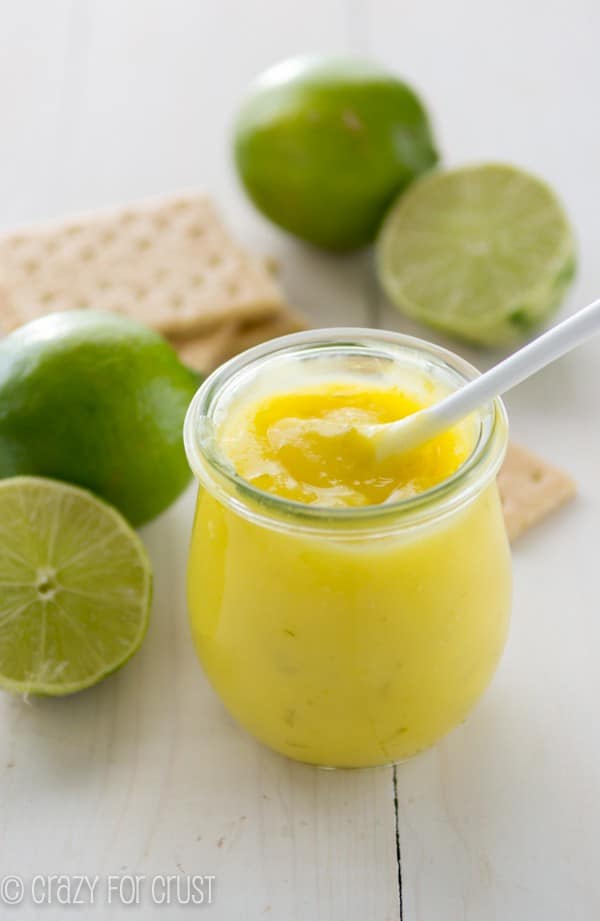 Homemade Lime Curd in a small clear glass with limes and graham crackers spread all around 