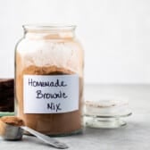 A large glass jar labeled Homemade Brownie Mix sits on a counter, filled with homemade brownie mix. A metal measuring spoon of mix and a plate with brownies are nearby. The jar’s lid rests beside it.