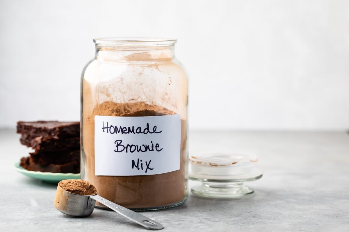 A large glass jar labeled Homemade Brownie Mix sits on a counter, filled with homemade brownie mix. A metal measuring spoon of mix and a plate with brownies are nearby. The jar’s lid rests beside it.