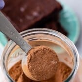 A tablespoon measuring spoon filled with cocoa powder is held over an open jar of cocoa powder, perfect for preparing homemade brownie mix, with two pieces of chocolate cake on a plate in the blurred background.