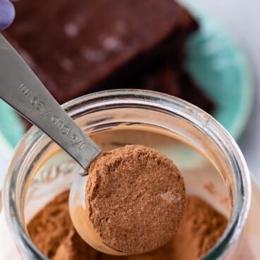 A tablespoon measuring spoon filled with cocoa powder is held over an open jar of cocoa powder, perfect for preparing homemade brownie mix, with two pieces of chocolate cake on a plate in the blurred background.