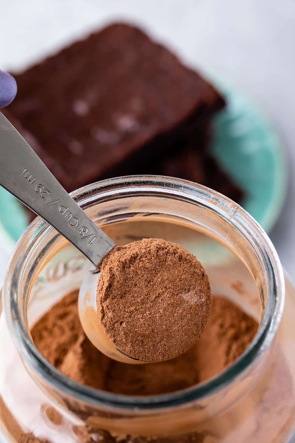 A tablespoon measuring spoon filled with cocoa powder is held over an open jar of cocoa powder, perfect for preparing homemade brownie mix, with two pieces of chocolate cake on a plate in the blurred background.