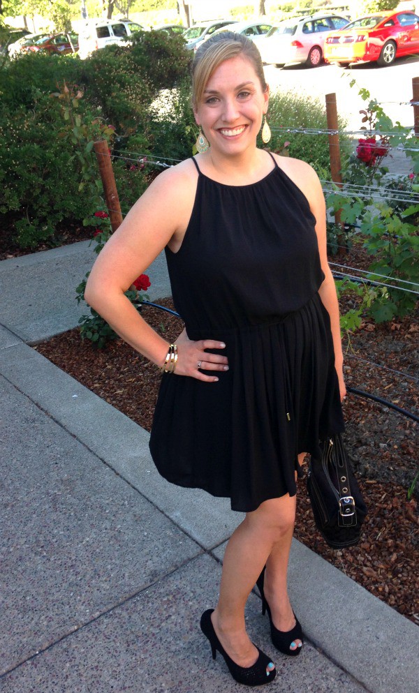 woman standing, wearing a black dress holding a black purse with black high heels with her hand on her hip