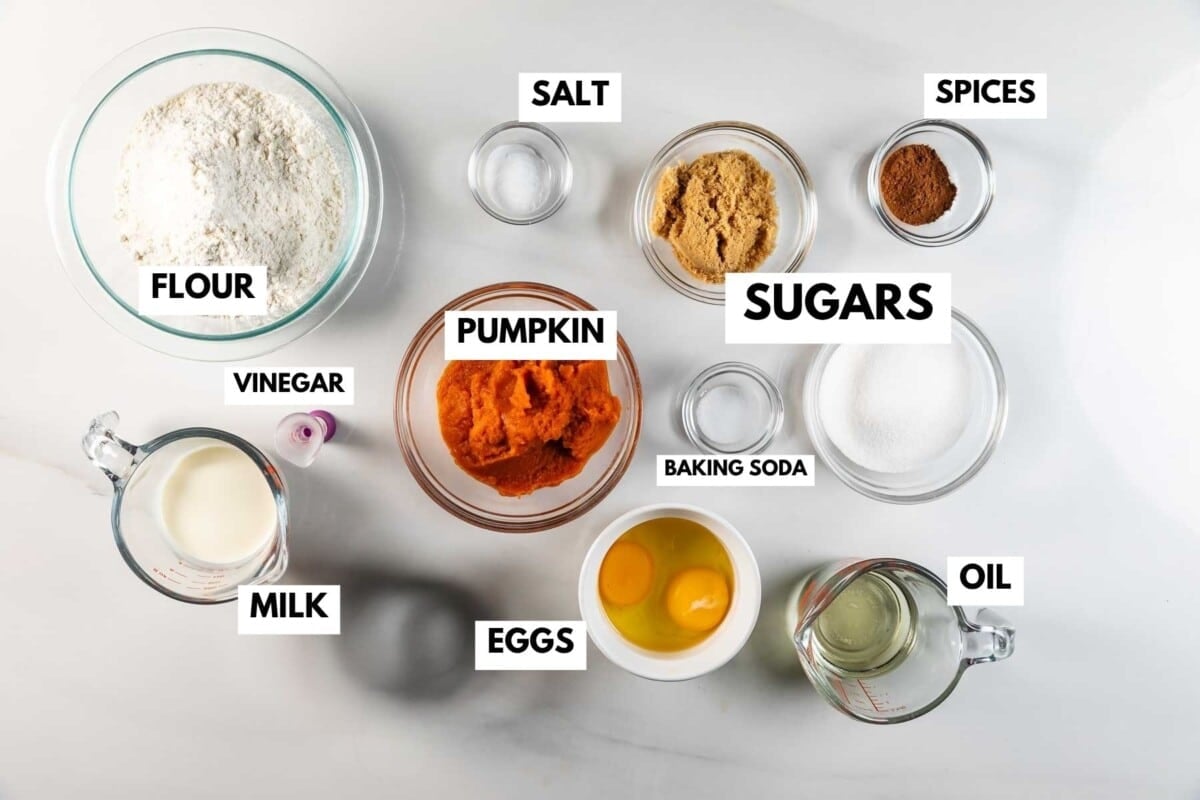 Top-down view of labeled baking ingredients in glass bowls for pumpkin bread with maple glaze: flour, pumpkin puree, eggs, milk, oil, sugars, brown sugar, spices, salt, vinegar, and baking soda on a white surface.