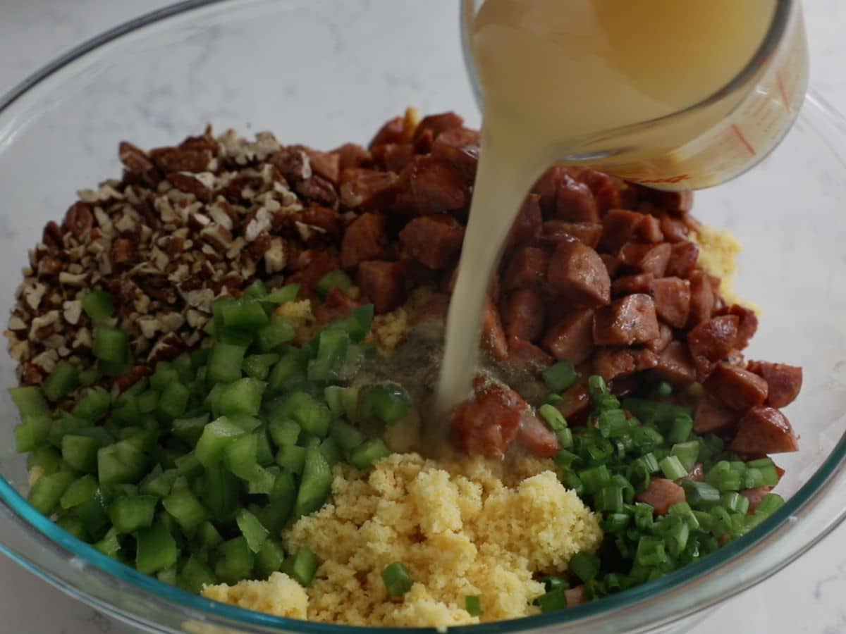 crumbled cornbread with sausage, pecans, green peppers, and green onions in large glass bowl with measuring cup pouring in chicken stock.