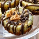 Close-up of several Chocolate Chip Cookie Cheesecakes topped with chocolate chips and cookie crumbles, finished with a decorative chocolate drizzle, arranged on a glass stand.