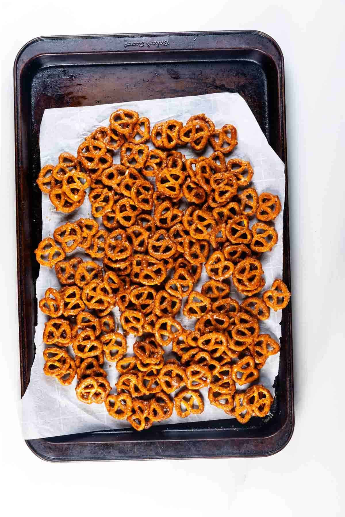 A baking sheet lined with parchment paper holds a single layer of sweet and spicy pretzels, spread evenly across the surface. The pretzels appear crisp and golden brown.