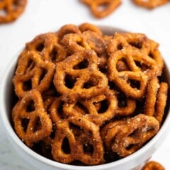 A white bowl filled with crunchy, sweet and spicy pretzels sits on a white surface, with more pretzels scattered in the background.
