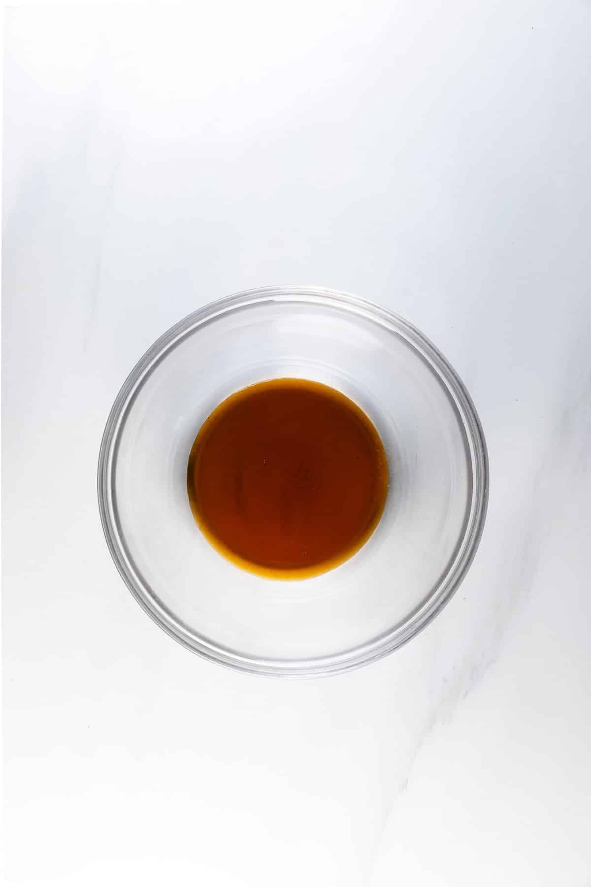 A clear glass bowl filled with a small amount of brown liquid, placed on a white marble surface—perfect for dipping sweet and spicy pretzels.