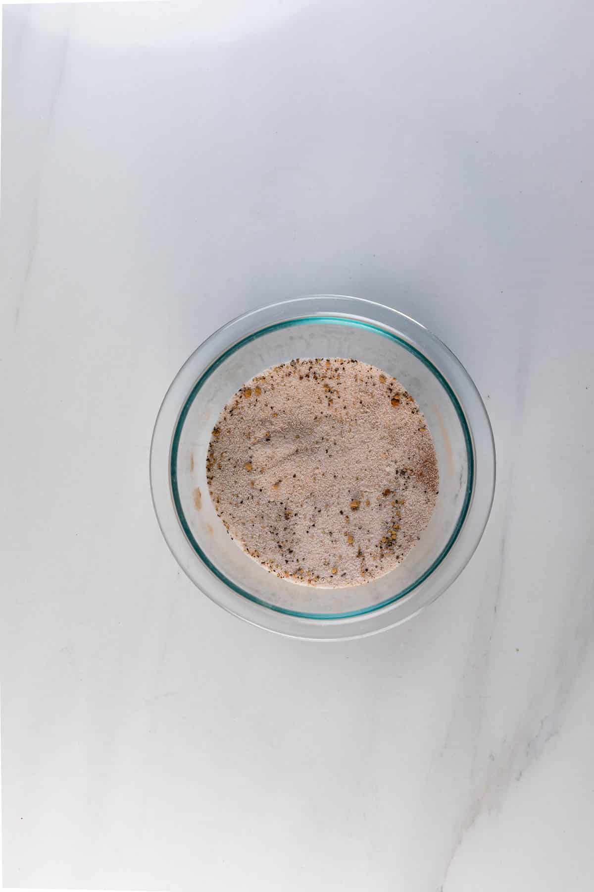 A glass bowl filled with a light brown, speckled powder mixture—perfect for coating sweet and spicy pretzels—sits on a white marble surface.