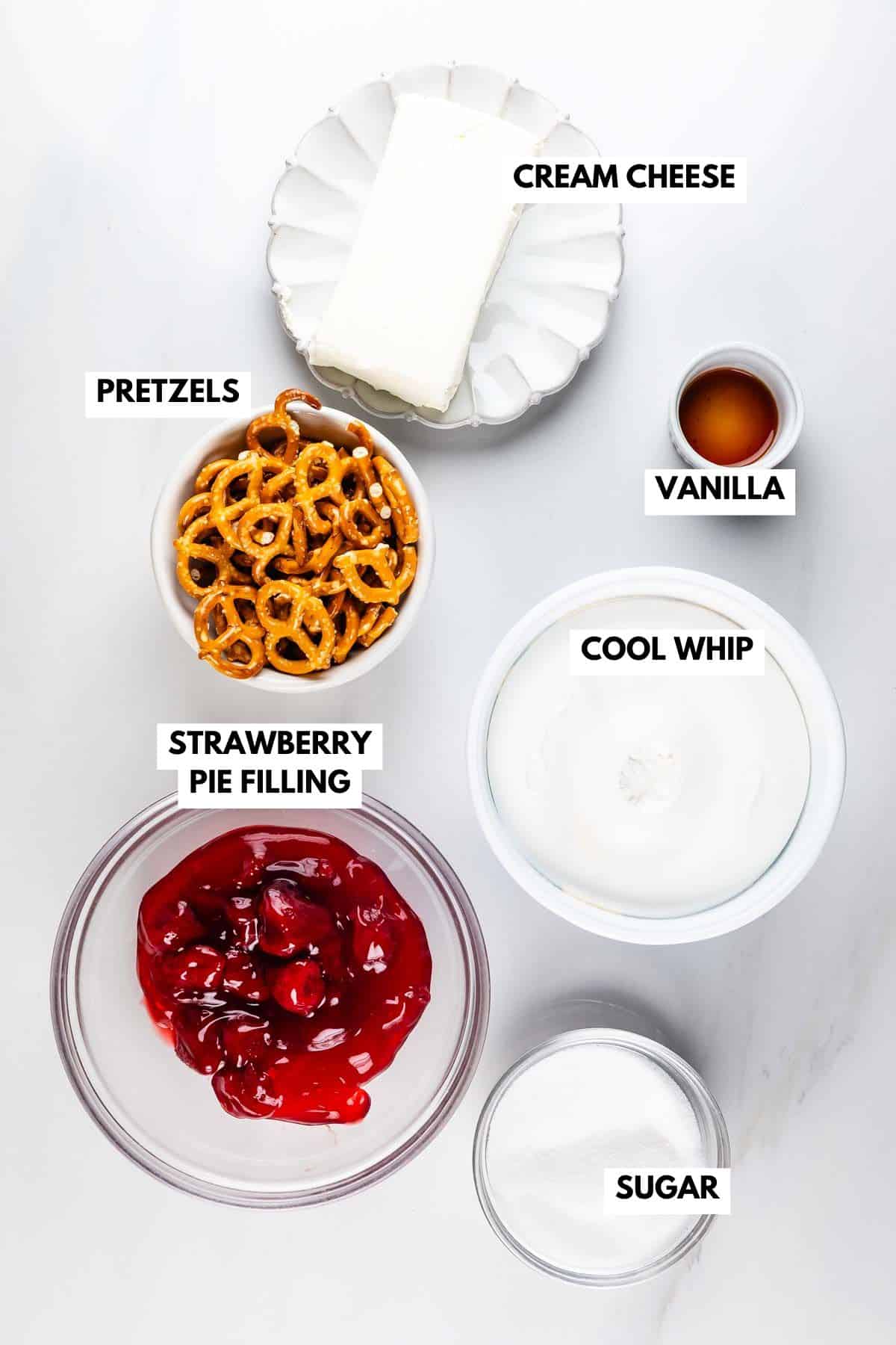 A photo showcases ingredients for a Strawberry Pretzel Salad Dip: a plate of cream cheese, a bowl of pretzels, a dish of vanilla extract, a tub of Cool Whip, strawberry pie filling, and sugar, all neatly labeled.