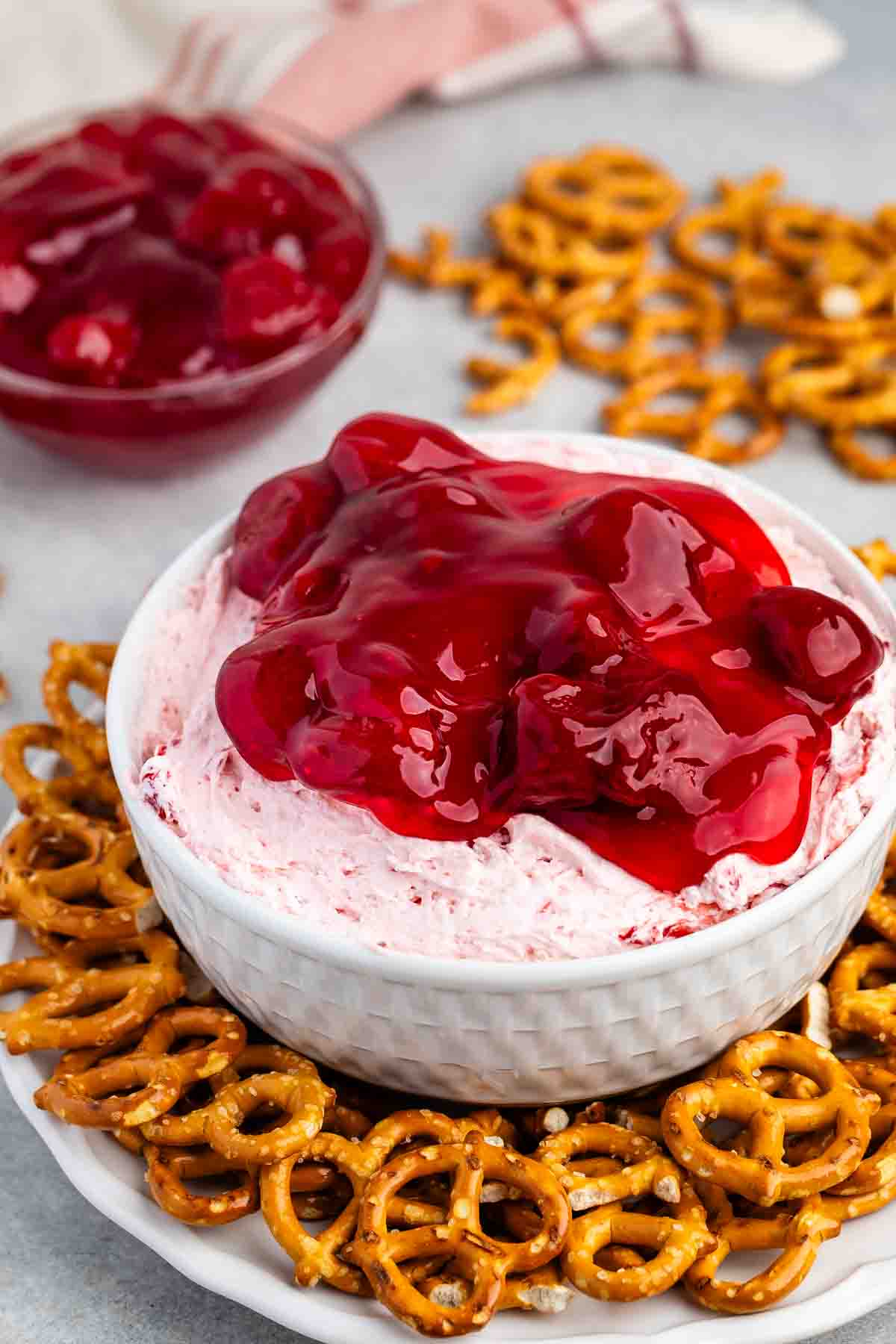 A white bowl filled with creamy pink Strawberry Pretzel Salad Dip topped with cherry sauce sits on a plate surrounded by mini pretzels. In the background, theres a small bowl of cherry sauce and a few scattered pretzels on a light-colored surface.