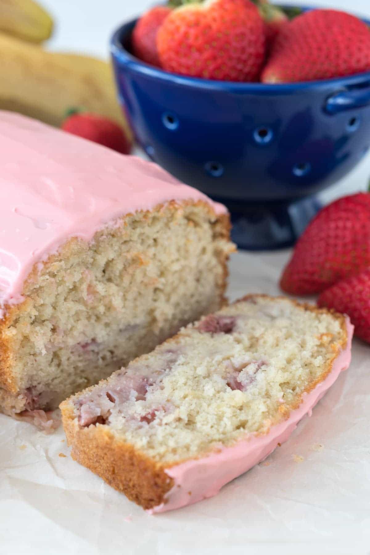 Sliced Strawberry Banana Bread on paper with a blue bowl full of strawberries. 