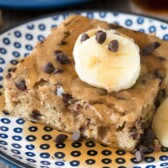 Slice of banana chocolate chip pancake cake on blue and white plate drizzled in syrup with bananas on top