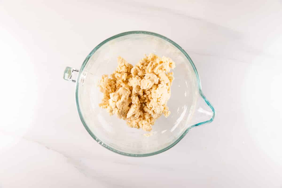 A glass mixing bowl with a handle holds a mound of light, crumbly dough mixture for irresistible Orange Dream Bars, resting on a white marble surface.