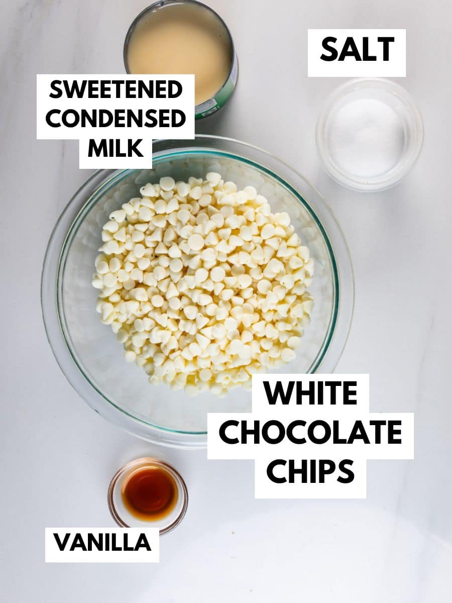 A glass bowl filled with white chocolate chips sits on a white surface, surrounded by small bowls of sweetened condensed milk, salt, and vanilla—everything you need for easy 5 Minute White Chocolate Fudge. Each ingredient is clearly labeled with bold text.