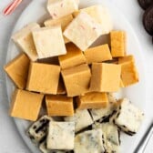 An oval white plate displays 5 Minute White Chocolate Fudge in three varieties: white fudge with pink peppermint pieces, light brown peanut butter fudge, and cookies and cream. Candy canes, white chocolate chips, and chocolate cookies are nearby.