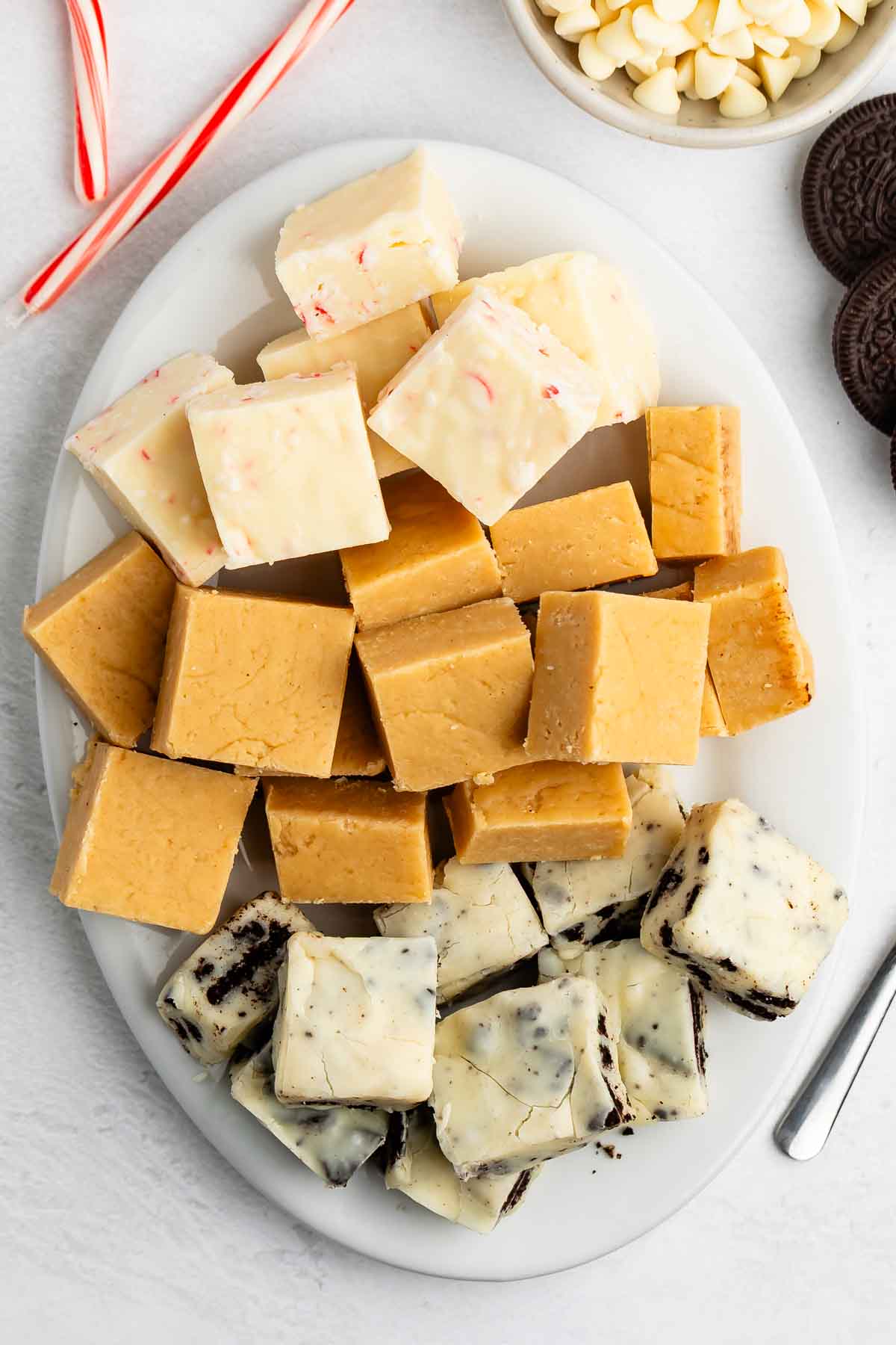 An oval white plate displays 5 Minute White Chocolate Fudge in three varieties: white fudge with pink peppermint pieces, light brown peanut butter fudge, and cookies and cream. Candy canes, white chocolate chips, and chocolate cookies are nearby.