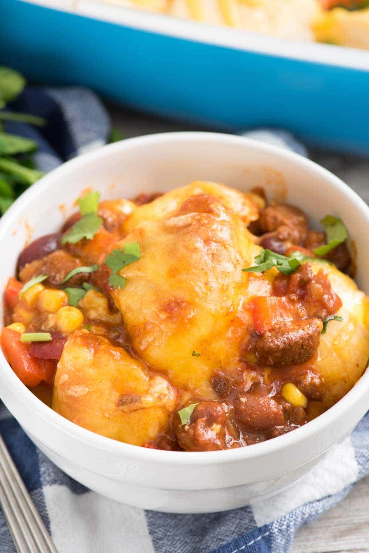 Easy Cheesy Chili Biscuit Bakein a white bowl - this easy weeknight dinner recipe has just 5 ingredients! Chili is topped with vegetables, biscuits, and cheese then baked up for a delicious comfort food meal.